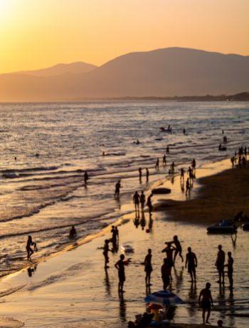 Spiaggia al tramonto con persone che camminano e nuotano, montagne sullo sfondo.