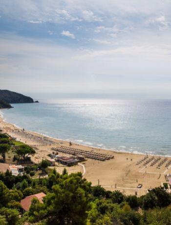 Spiaggia sabbiosa con ombrelloni, mare calmo e colline verdi sullo sfondo.
