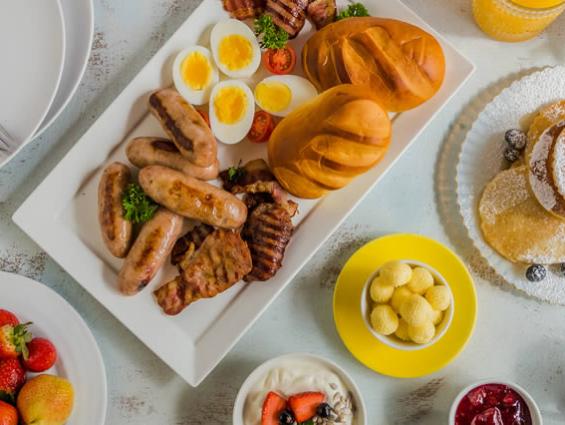 Colazione abbondante con salsicce, uova, pane, frutta, pancake e yogurt.