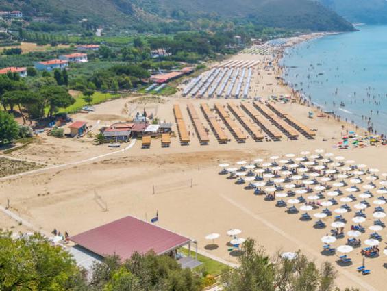 Spiaggia affollata con ombrelloni, mare calmo e colline sullo sfondo.