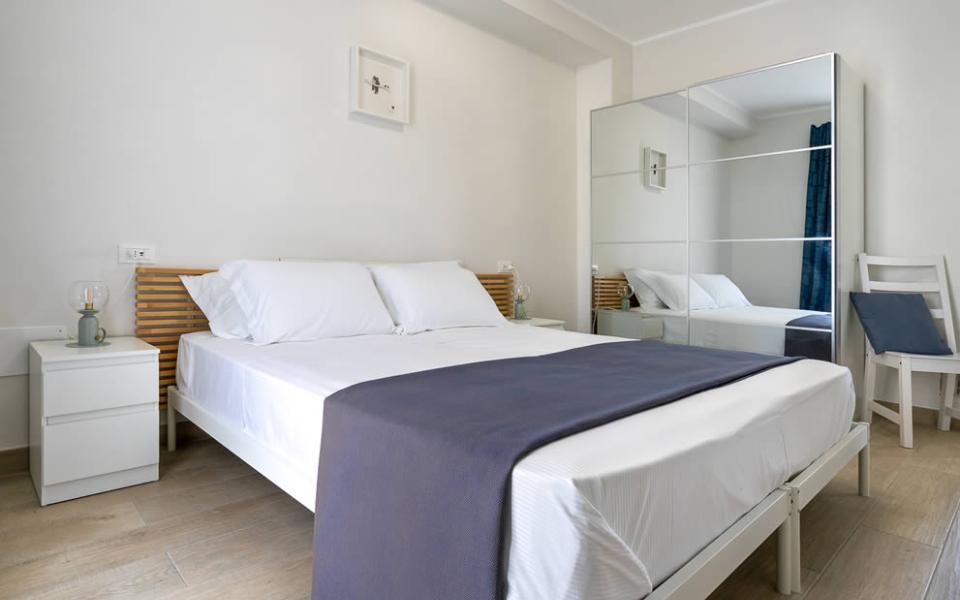 Camera moderna e luminosa, letto bianco con coperta grigia, atmosfera serena e accogliente.