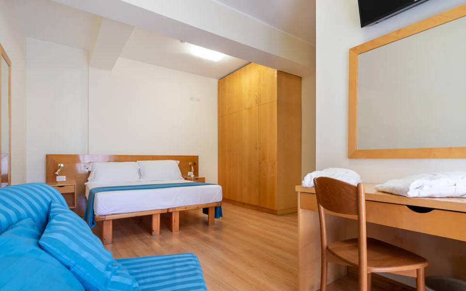 Camera accogliente con letto matrimoniale, divano azzurro, mobili in legno chiaro, atmosfera rilassante.