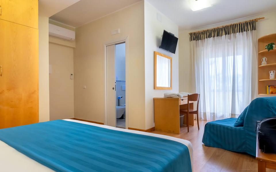 Camera luminosa, accogliente, con letto blu, scrivania, TV e bagno privato.
